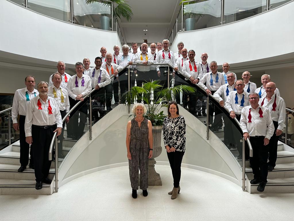 Photo of all the choir members on a staircase with MD Jane & accompanist Kerry-Anna standing infront